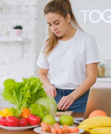 Woman in white t-shirt preparing fresh vegetables in a bright kitchen with a laptop.