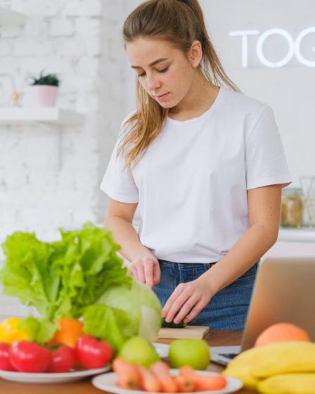 Woman in white t-shirt preparing fresh vegetables in a bright kitchen with a laptop.