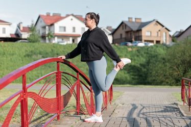 A woman in active wear performs a stretching exercise on a red bridge in a suburban park setting.