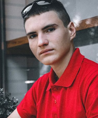 Portrait of a young man wearing a red polo shirt indoors, looking directly at the camera.
